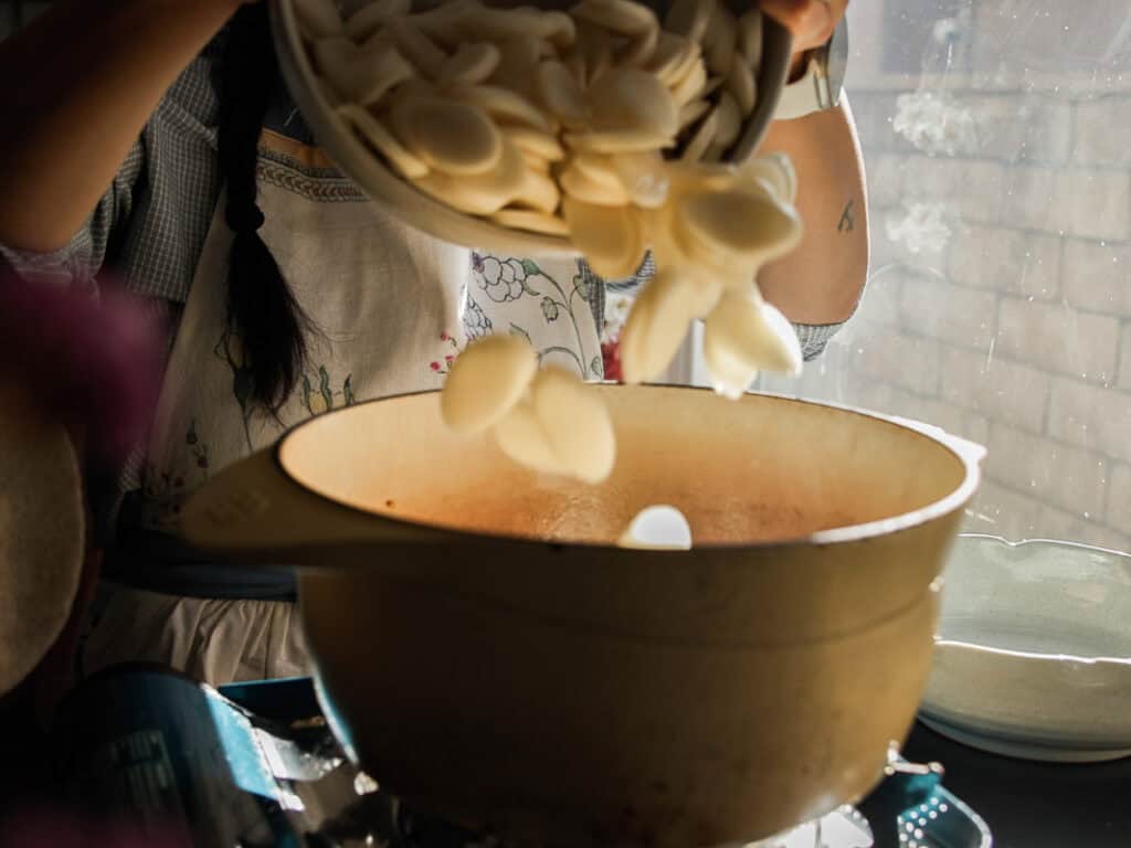 A person wearing an apron pours sliced rice cakes from a bowl into a pot on a stove, with sunlight streaming through a window beside them.